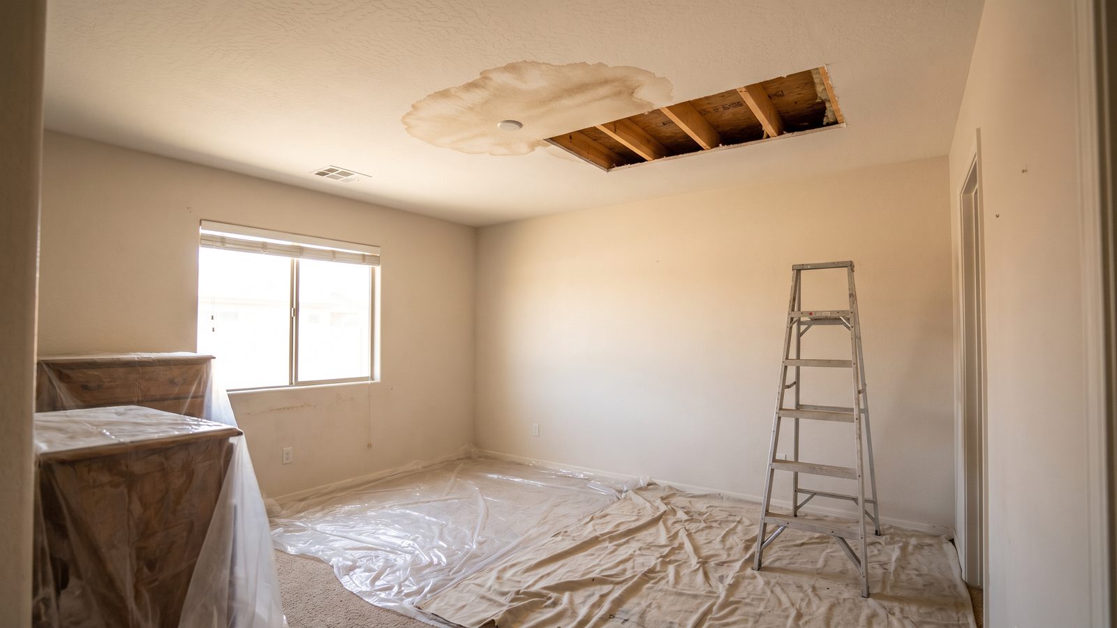 A brown water stain spread across a bedroom ceiling with a rectangular section of ceiling board removed for drying, exposing the joists behind, and plastic sheeting and a step ladder set up below