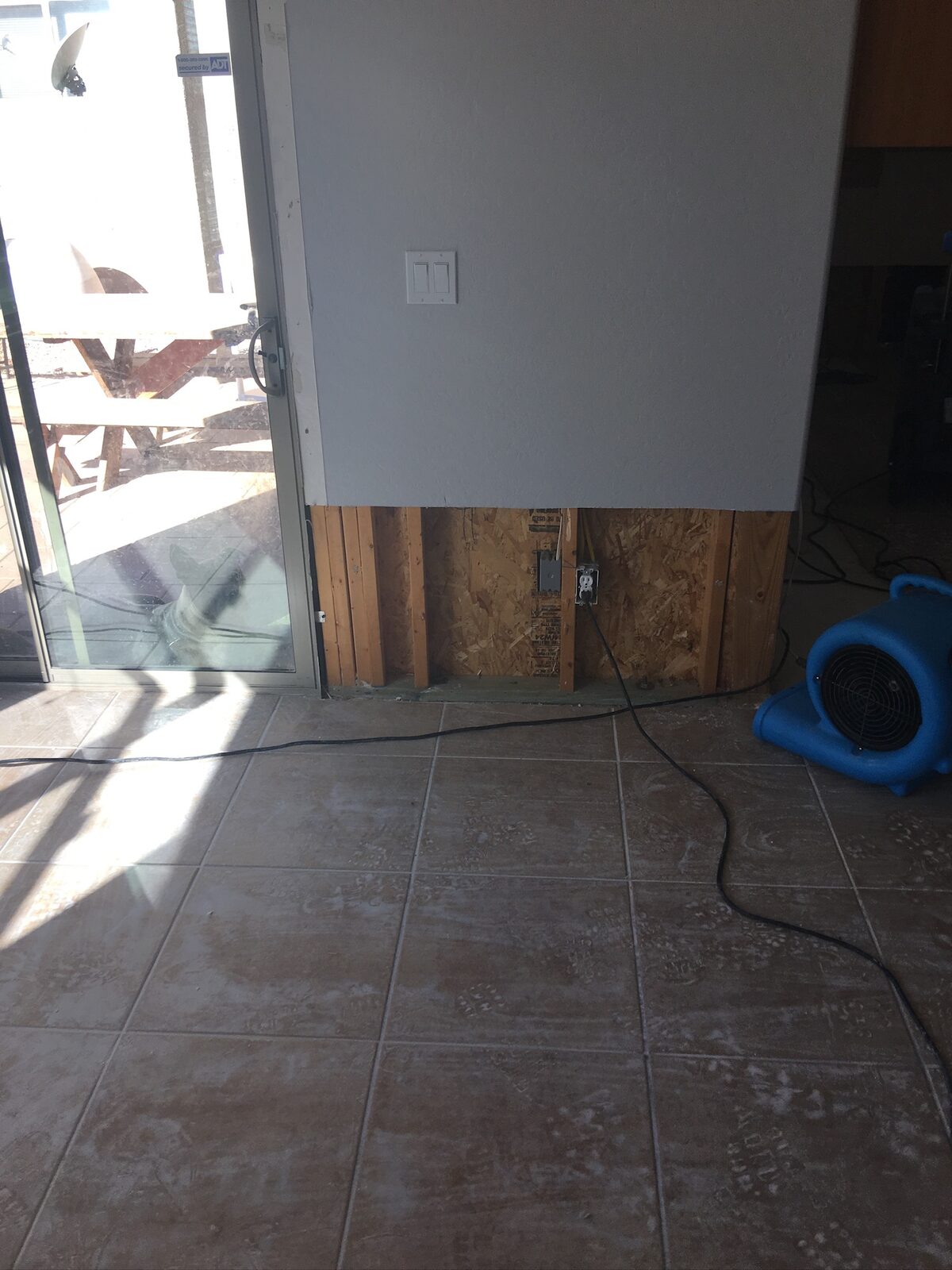 Air mover beside a wall with lower drywall removed to expose sheathing near a sliding door