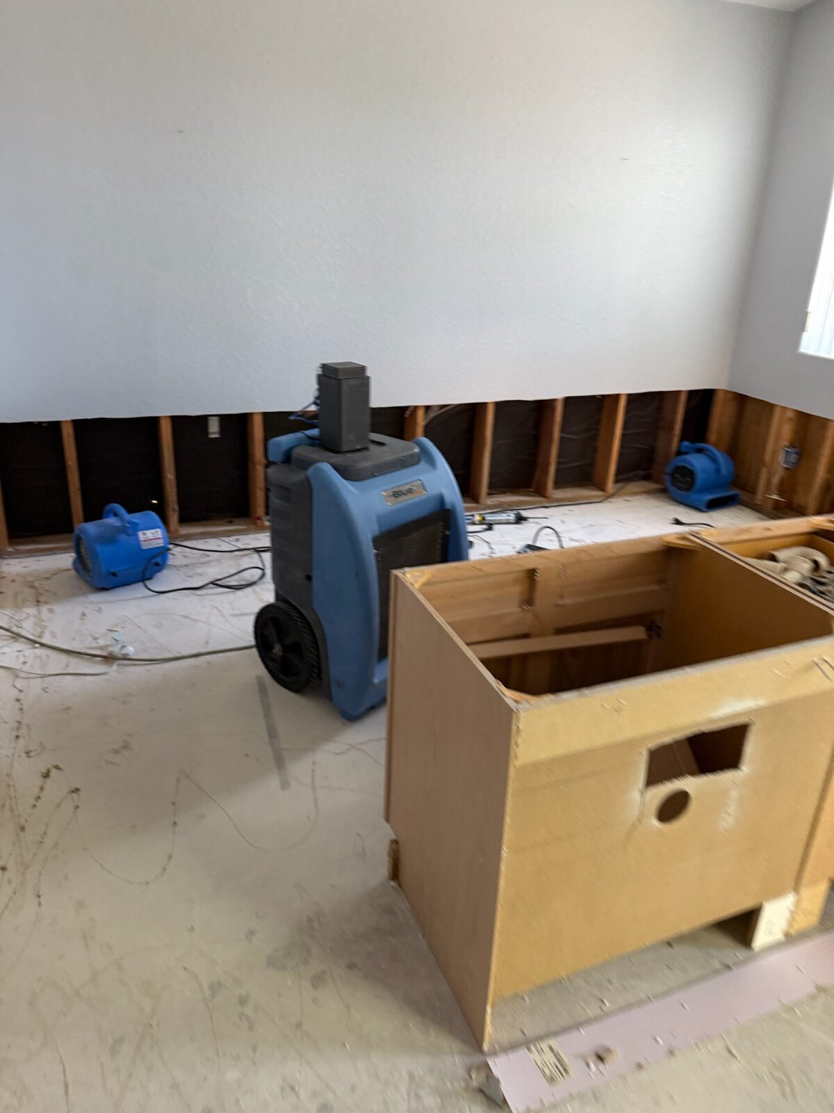 Dehumidifier and air movers running beside a cabinet box in a room with flood-cut drywall