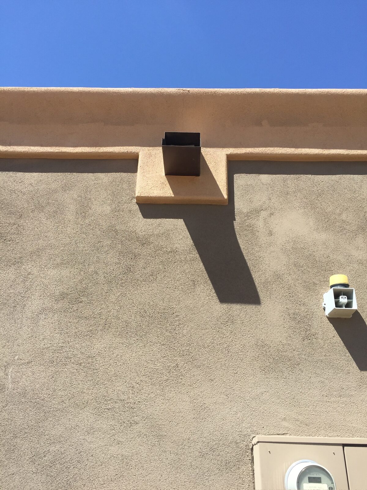 Stucco parapet wall with a metal scupper drain opening under a clear sky
