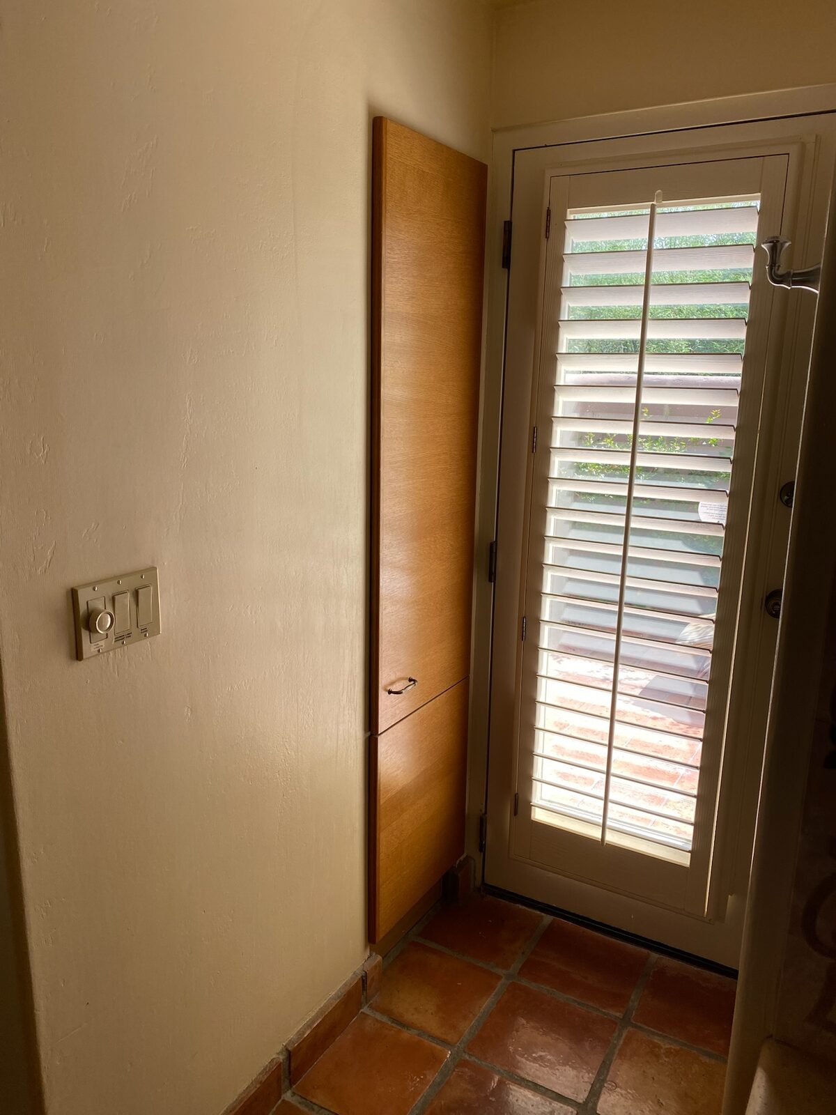 Tall wood cabinet beside a glass door with plantation shutters and terracotta tile floor