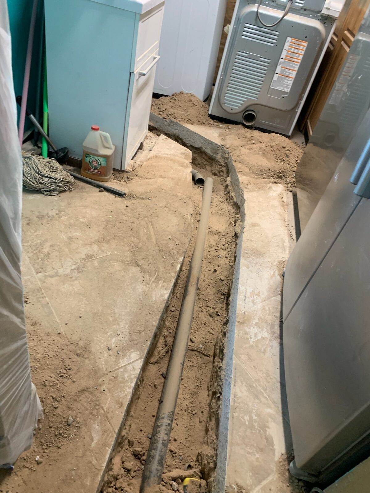 Trench cut through a concrete slab with a new drain line routed past laundry appliances